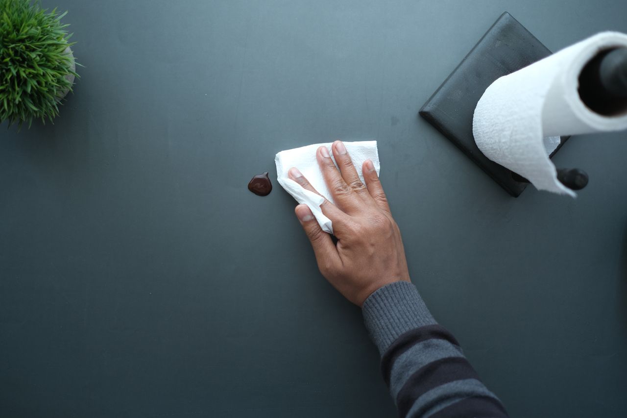 Une main essuie une surface sombre avec un mouchoir, près d’un rouleau de papier et d’une plante.